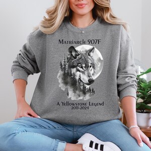 May include: Gray sweatshirt with a black and white wolf illustration, a full moon, and trees. The text "Matriarch 907F A Yellowstone Legend 2015-2024" is printed on the sweatshirt.