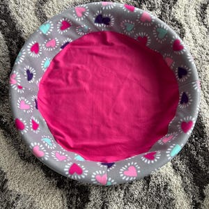 May include: A round pet bed with a gray exterior patterned with pink, purple, and teal hearts. The interior is a solid bright pink. The bed is designed for small pets.