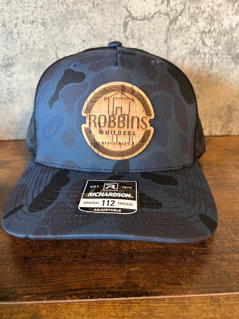 Op de afbeelding: Een blauwe en zwarte camouflage trucker-pet met een bruine houten patch waarop "Robbins Builders" en "Spreuken 22:29" staat. De pet heeft een verstelbare band en een Richardson-logo.