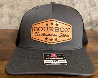 May include: A gray and black trucker hat with a brown leather patch that says "Bourbon The American Spirit" with five stars. The hat is adjustable and has the Richardson 112 logo on the side.