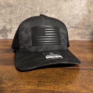 Può includere: Cappellino da baseball mimetico nero con una toppa nera con la bandiera americana. Il cappellino ha un retro in rete e una visiera curva. L'etichetta recita "Richardson 112 Original Trucker Adjustable".