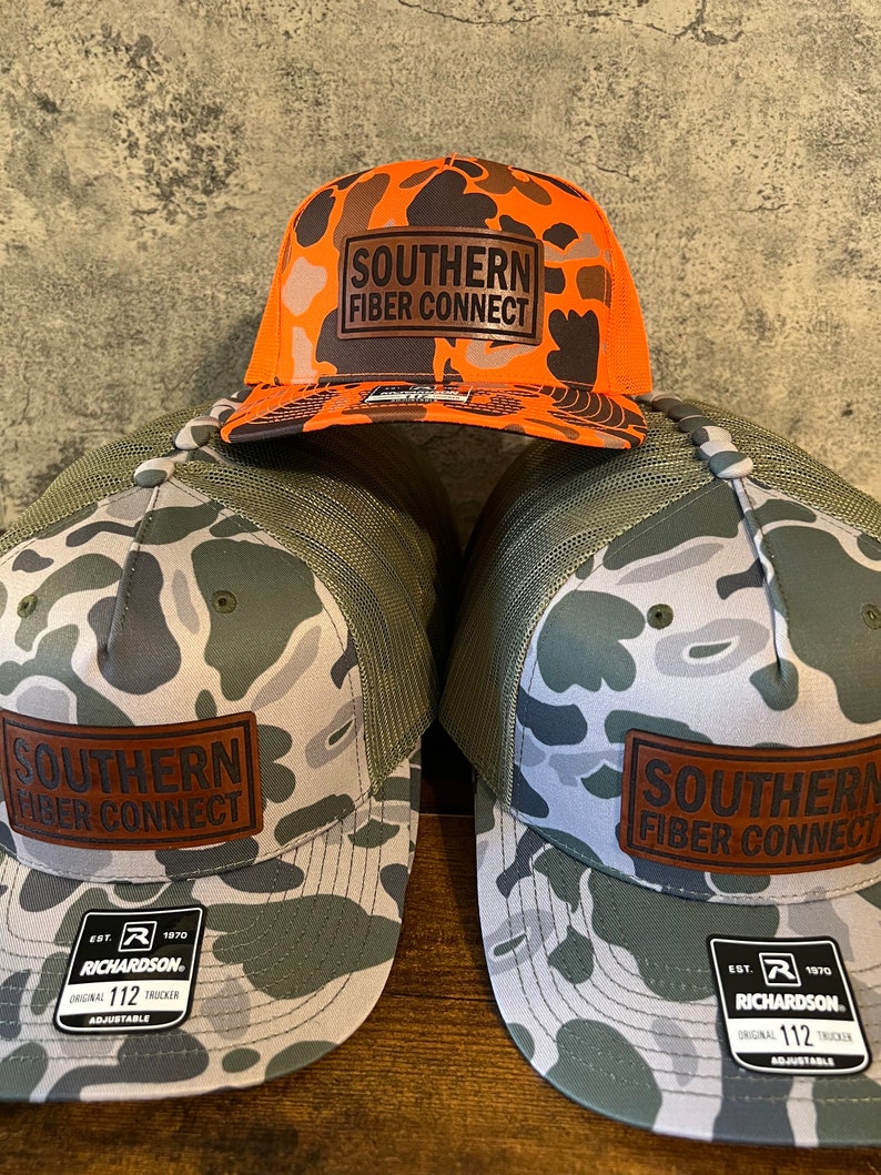 Op de afbeelding: Drie camouflage trucker caps met een bruin leren patch waarop "Southern Fiber Connect" staat. De caps zijn allemaal in verschillende camouflagekleuren, een is oranje, een is groen en een is grijs.
