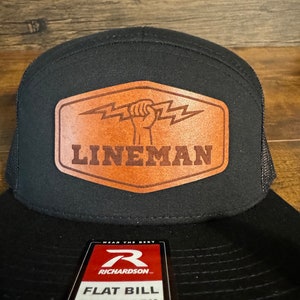 Può includere: Cappellino nero e marrone con visiera piatta e chiusura a pressione con una toppa in pelle che dice "LINEMAN" e raffigura un pugno con un fulmine. Il cappello ha anche un'etichetta rossa e nera che dice "WEAR THE BEST RICHARDSON FLAT BILL SNAPBACK EST 1979".