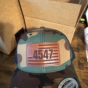 May include: A camouflage trucker hat with a brown leather patch featuring the number "4547" and a small American flag. The hat has a black mesh back and a black and green camouflage front.