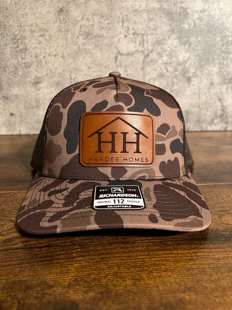 Op de afbeelding: Een bruine en zwarte camouflage trucker-pet met een bruin leren patch waarop "HH Hardee Homes" staat. De pet heeft een zwart mesh-achterpaneel en een verstelbare band met een zwart label waarop "Richardson 112 Original Trucker Adjustable" staat.