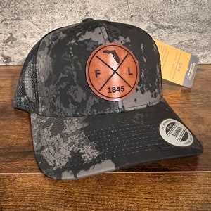 May include: Black and gray camouflage trucker hat with a brown leather patch featuring the outline of Florida and the letters "F" and "L" with a cross in between and the year "1845" below. The hat has a black mesh back.