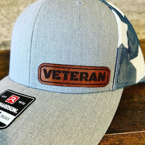 May include: A grey and white trucker hat with a brown leather patch that says "Veteran". The hat has a mesh back and a Richardson logo on the side.