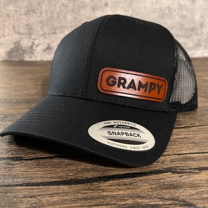 May include: Black mesh and twill baseball cap with a brown leather patch that says "GRAMPY" and a white oval sticker that says "The Authentic Yupoong Snapback Yupoong Since 1974".