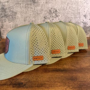 May include: A row of five baseball caps. The caps have a light blue front panel and a khaki-colored brim and back. The caps have a perforated mesh back panel. Each cap has a rectangular patch with the letters "HN UT".