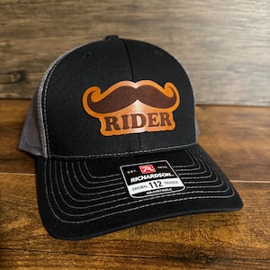 Puede incluir: Gorra de camionero negra y gris con un parche de cuero marrón que dice "RIDER" con un bigote marrón sobre el texto. La gorra tiene una correa ajustable negra con una etiqueta blanca que dice "EST. 1970 RICHARDSON ORIGINAL 112 TRUCKER ADJUSTABLE".