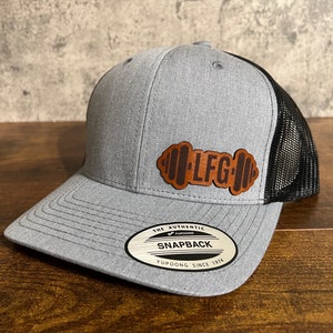 May include: A grey and black mesh trucker hat with a brown leather patch that says "LFG" in a stylised font. The hat has a YUPOONG label that says "The Authentic Snapback YUPOONG Since 1974".
