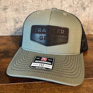 Könnte beinhalten: Eine grüne und schwarze Trucker-Kappe mit einem schwarzen Leder-Patch, auf dem "Tranter Grey" steht, und einem roten und schwarzen Etikett, auf dem "Richardson 112 Original Trucker Adjustable" steht.