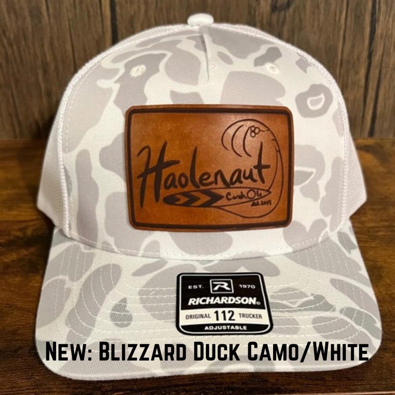 Op de afbeelding: Witte en grijze camouflage trucker cap met een bruin leren patch waarop "Haolenaut" staat en een wit en grijs Richardson label waarop "EST. 1970 ORIGINAL 112 TRUCKER ADJUSTABLE" staat.
