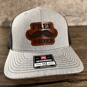 Puede incluir: Una gorra de camionero gris y negra con un parche de cuero marrón que dice "FREE RIDES" con un bigote negro sobre el texto. La gorra es ajustable y tiene una etiqueta Richardson 112.