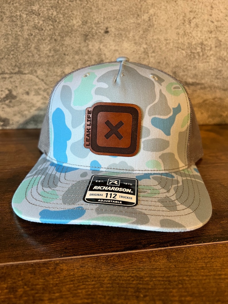 Op de afbeelding: Een grijze, blauwe en groene camouflage trucker-pet met een bruine leren patch met een witte 'X' en de tekst 'LEAK LIFE'. De pet heeft een zwart mesh-rug en een zwarte verstelbare band met een zwarte plastic gesp. De pet heeft ook een zwart label met de tekst 'EST. 1970 RICHARDSON ORIGINAL 112 TRUCKER ADJUSTABLE'.