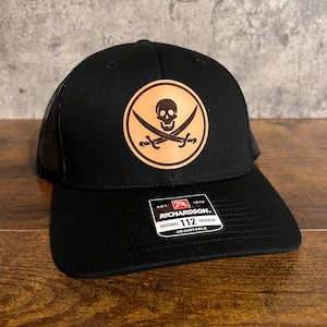 May include: Black trucker hat with a mesh back and a curved brim. The hat features a circular patch with a skull and crossed swords design. The tag reads "RICHARDSON ORIGINAL 112 TRUCKER ADJUSTABLE".
