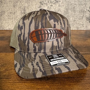 Peut inclure: Casquette de camionneur camouflage avec un écusson en forme de plume marron et blanche. Le chapeau a un dos en filet et une visière incurvée. L'étiquette indique "RICHARDSON 112 TRUCKER ADJUSTABLE".