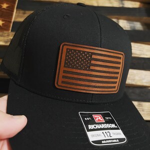 May include: Black trucker hat with a brown leather American flag patch and a black and red Richardson 112 adjustable label.