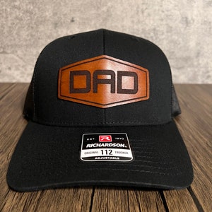 May include: Black trucker hat with a brown leather patch that says "DAD" on it. The hat is adjustable and has a Richardson 112 label.