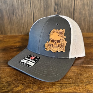May include: A gray and white trucker hat with a brown leather patch featuring a skull with roses. The patch is attached to the front of the hat. The hat has a Richardson logo on the side.