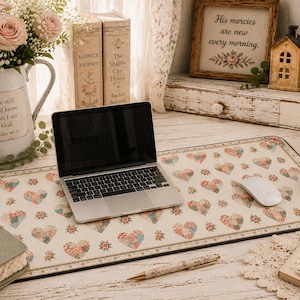 May include: A rectangular desk mat featuring a repeating pattern of multicolored floral hearts and small star-like motifs on a cream background. The mat has a dark border and is positioned under a silver laptop and a white computer mouse.
