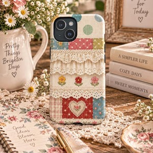 Puede incluir: Una funda para smartphone con un diseño de patchwork que incluye lunares, motivos florales y un corazón central. La funda está decorada con capas horizontales de encaje blanco con ojales, creando un aspecto texturizado y superpuesto.