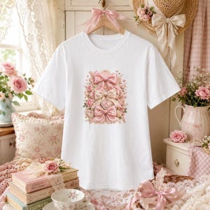 Könnte beinhalten: Ein weißes T-Shirt mit kurzen Ärmeln und einem zentralen Grafikdruck aus drei übereinanderliegenden rosa Schleifen, umgeben von zarten rosa Rosen und floralen Akzenten. Das T-Shirt hat einen Rundhalsausschnitt und hängt an einem Kleiderbügel mit rosa Band.