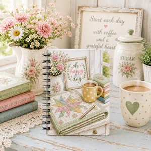 May include: A spiral-bound notebook featuring a printed illustration of a cozy scene with a quilt, a yellow polka-dot mug, and a framed sign that says "oh happy day." The notebook cover displays a soft color palette with floral and quilted patterns.