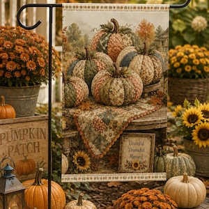 May include: A rectangular garden flag featuring a printed scene of quilted fabric pumpkins in shades of green, orange, and tan resting on a wooden table. A small sign at the bottom reads Grateful, Thankful, Blessed with a sunflower graphic.