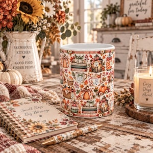 May include: A white ceramic mug featuring a repeating autumn-themed pattern of pumpkins, stacked books, lanterns, and mugs with text including Cozy Nights, Sweet Treats, and Hello October, surrounded by fall foliage, berries, and small flowers.