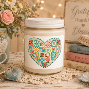 May include: A clear glass candle jar with a white lid features a label with a colorful heart-shaped quilt pattern. The heart design is composed of various small squares in shades of pink, blue, yellow, and green, surrounded by a decorative lace border.