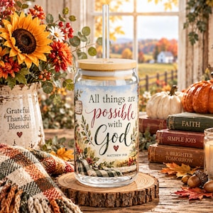 May include: A clear glass tumbler with a bamboo lid and a glass straw, featuring a printed design that reads All things are possible with God and Matthew 19:26, surrounded by colorful autumn foliage and a small red heart illustration.