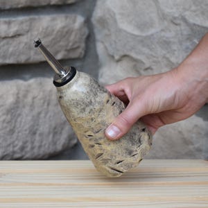 May include: A ceramic oil cruet with a textured, speckled beige and grey finish. The bottle features a tapered body with carved indentations and is topped with a stainless steel pour spout featuring a black rubber seal for secure closure.
