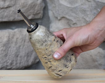 May include: A ceramic oil cruet with a textured, speckled beige and grey finish. The bottle features a tapered body with carved indentations and is topped with a stainless steel pour spout featuring a black rubber seal for secure closure.
