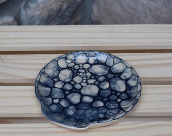 May include: A small, round ceramic dish featuring a distinctive bubble-like pattern in shades of dark blue and off-white. The surface displays a dense, interconnected network of circular shapes that vary in size, creating a textured appearance.
