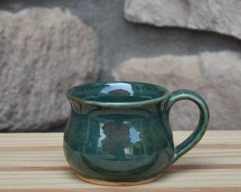 May include: A rounded ceramic mug with a glossy, deep teal and forest green glaze. The mug features a wide, curved body, a flared rim, and a sturdy, integrated handle on the right side. The surface has subtle variations in color and tone.