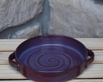 May include: A round, deep purple ceramic baking dish with two side handles. The interior surface features a prominent, hand-thrown spiral pattern that radiates from the center toward the outer edges, finished with a glossy, smooth glaze.