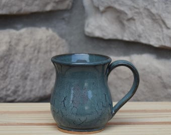 May include: A ceramic mug with a rounded body and a curved handle, featuring a glossy teal-blue glaze with a dark crackle pattern throughout the surface. The base of the mug reveals a natural, unglazed terracotta-colored clay rim.