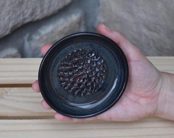 May include: A small, round, black ceramic grater plate with a glossy finish. The center features a raised, textured pattern designed for grating food, surrounded by a smooth, dark rim. The plate has a deep, reflective glaze.