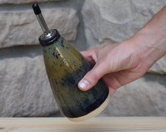 May include: A ceramic oil cruet with a tapered body featuring a mottled glaze in shades of olive green, brown, and deep blue. The vessel is topped with a stainless steel pourer spout that includes a small black plastic cap for sealing the opening.