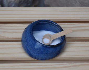 May include: A small, round, deep blue ceramic bowl with a glossy finish, filled with white granular salt. A small, light-colored wooden spoon rests inside the bowl, partially submerged in the salt, providing a contrast to the dark blue ceramic.