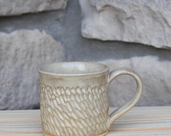 May include: A ceramic mug with a light beige, off-white glaze and a textured exterior featuring a repeating pattern of diagonal, carved indentations. The mug has a smooth, rounded handle and a cylindrical body with a slightly flared rim.