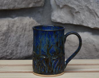 May include: A cylindrical ceramic mug featuring a deep blue and dark green glaze with a mottled, dripped appearance. The lower portion of the mug displays a textured, raised vertical pattern that wraps around the base, complemented by a curved handle.