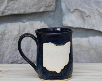 May include: A dark blue ceramic mug with a glossy finish featuring a cream-colored, unglazed cutout in the shape of the state of Ohio on the front. The mug has a curved handle on the left side and a cylindrical body with a flat base.
