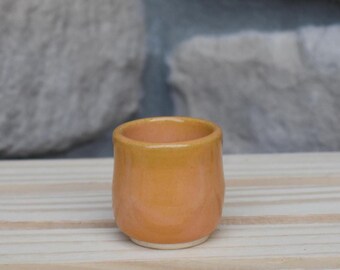 May include: A small, rounded ceramic cup with a smooth, glossy finish in a warm, vibrant orange hue. The vessel features a slightly tapered shape with a clean, unglazed white base rim, showcasing a simple, functional design for holding liquids.