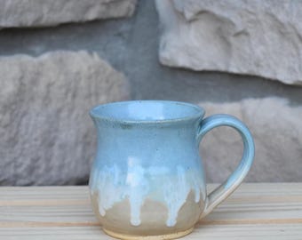 May include: A ceramic mug with a rounded body and a curved handle. The upper portion of the mug is a light blue color, while the lower portion is a tan color. A white glaze drips from the blue section down onto the tan base of the mug.