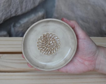 May include: A small, round, off-white ceramic dish with a smooth, glazed finish. The center of the dish features a detailed, embossed floral pattern consisting of numerous small, elongated petal shapes arranged in a circular, sunburst design.