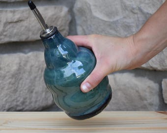 May include: A teal ceramic oil dispenser with a bulbous, wavy-textured body and a glossy finish. The vessel features a stainless steel pour spout with a black plastic cap at the top, designed for controlled pouring of liquid condiments or oils.