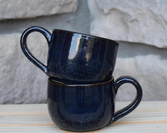 May include: Two stacked ceramic mugs with a glossy, deep navy blue glaze. Each mug features a rounded body, a curved handle on the side, and a smooth, reflective surface. The dark blue finish shows subtle variations in tone and texture throughout.