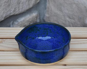 May include: A small, round, ceramic bowl with a deep blue glaze featuring mottled dark blue and black speckles. The bowl has a distinct, small pouring spout on one side and a smooth, light-colored base where the ceramic material is visible.